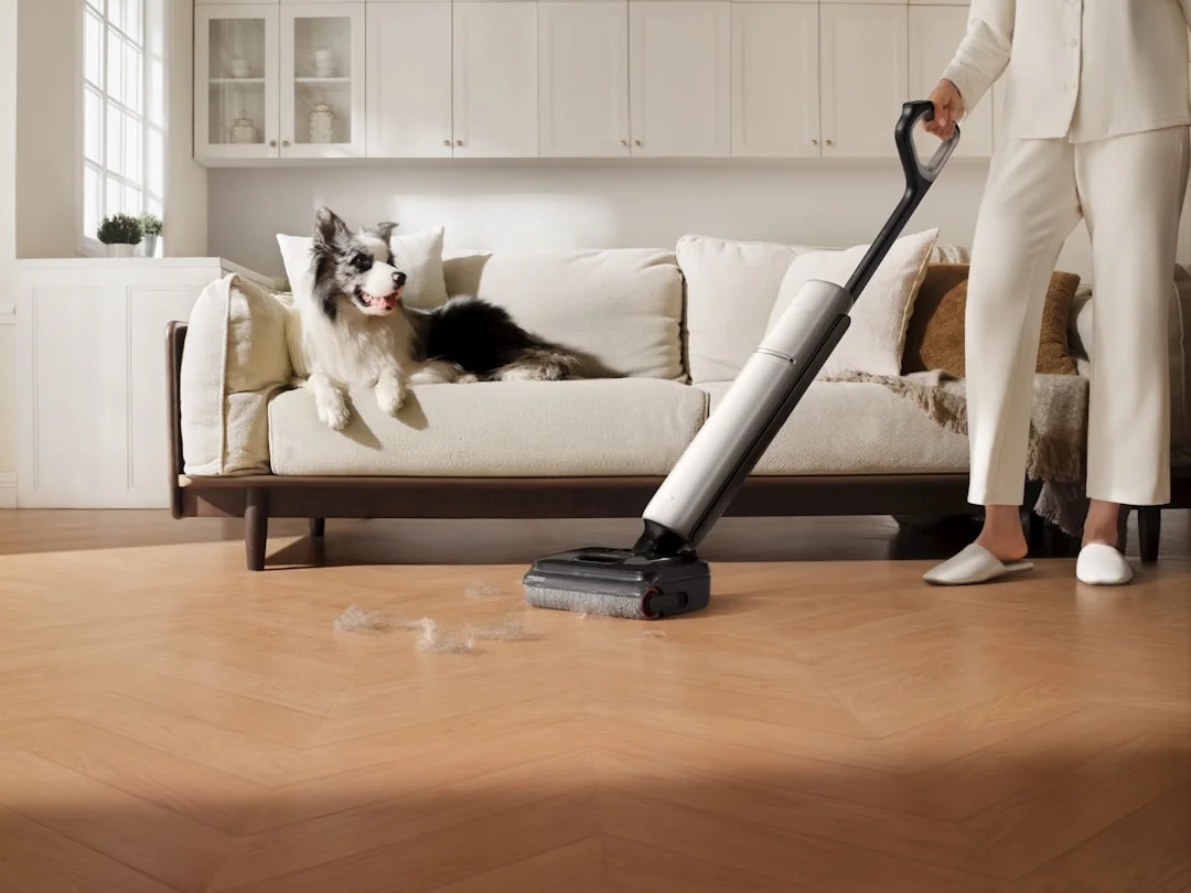 Two women in work clothes are cleaning a modern living room with a vacuum and a mop.